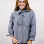 A smiling woman with long light brown hair wearing a blue and white vertically striped button-down shirt with a large bow tie collar, paired with dark blue jeans, standing against a white wood-paneled wall.