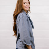 A woman with long light brown hair is shown from the side, wearing a blue and white vertically striped button-up shirt with a tie-neck detail and dark blue jeans, standing against a white shiplap wall.