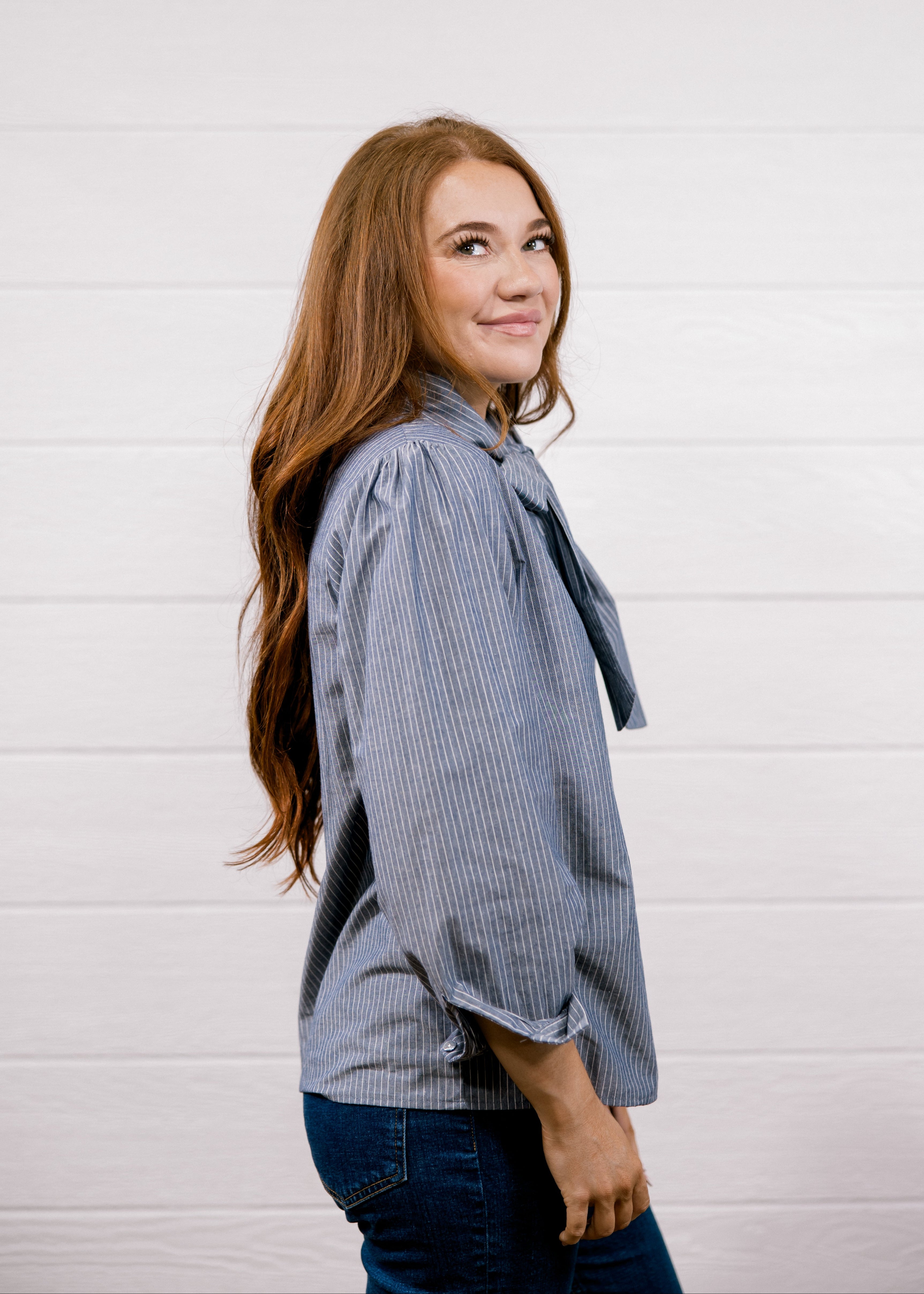 A woman with long light brown hair is shown from the side, wearing a blue and white vertically striped button-up shirt with a tie-neck detail and dark blue jeans, standing against a white shiplap wall.