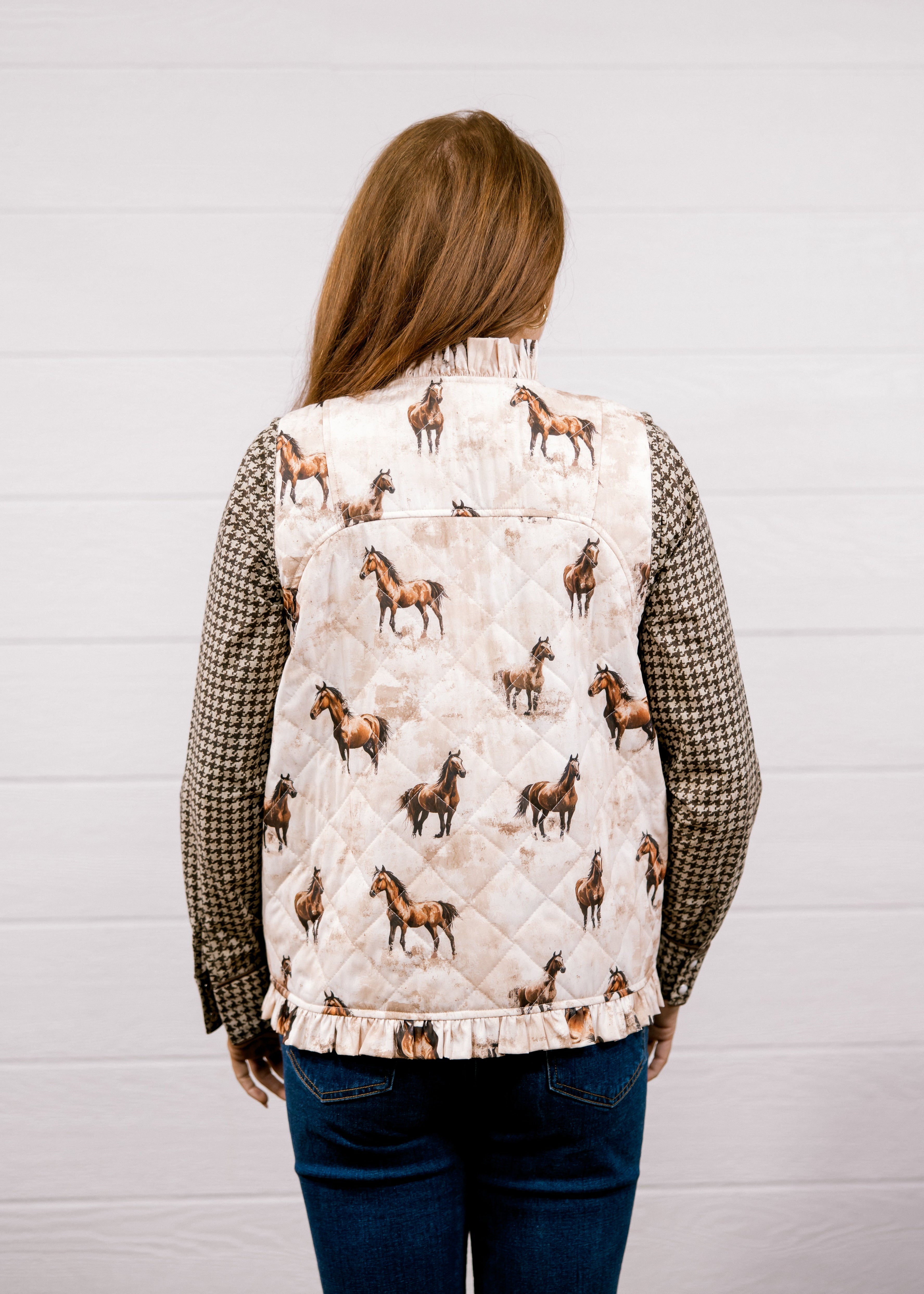 A woman viewed from behind wearing a quilted vest with a horse print on the back panel and checkered sleeves, paired with blue jeans, against a white wood-paneled wall.