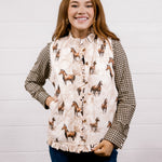 A smiling woman wearing a sleeveless western-style vest with a beige and brown horse print over a brown-and-white houndstooth shirt, paired with dark blue jeans, standing against a white shiplap wall.