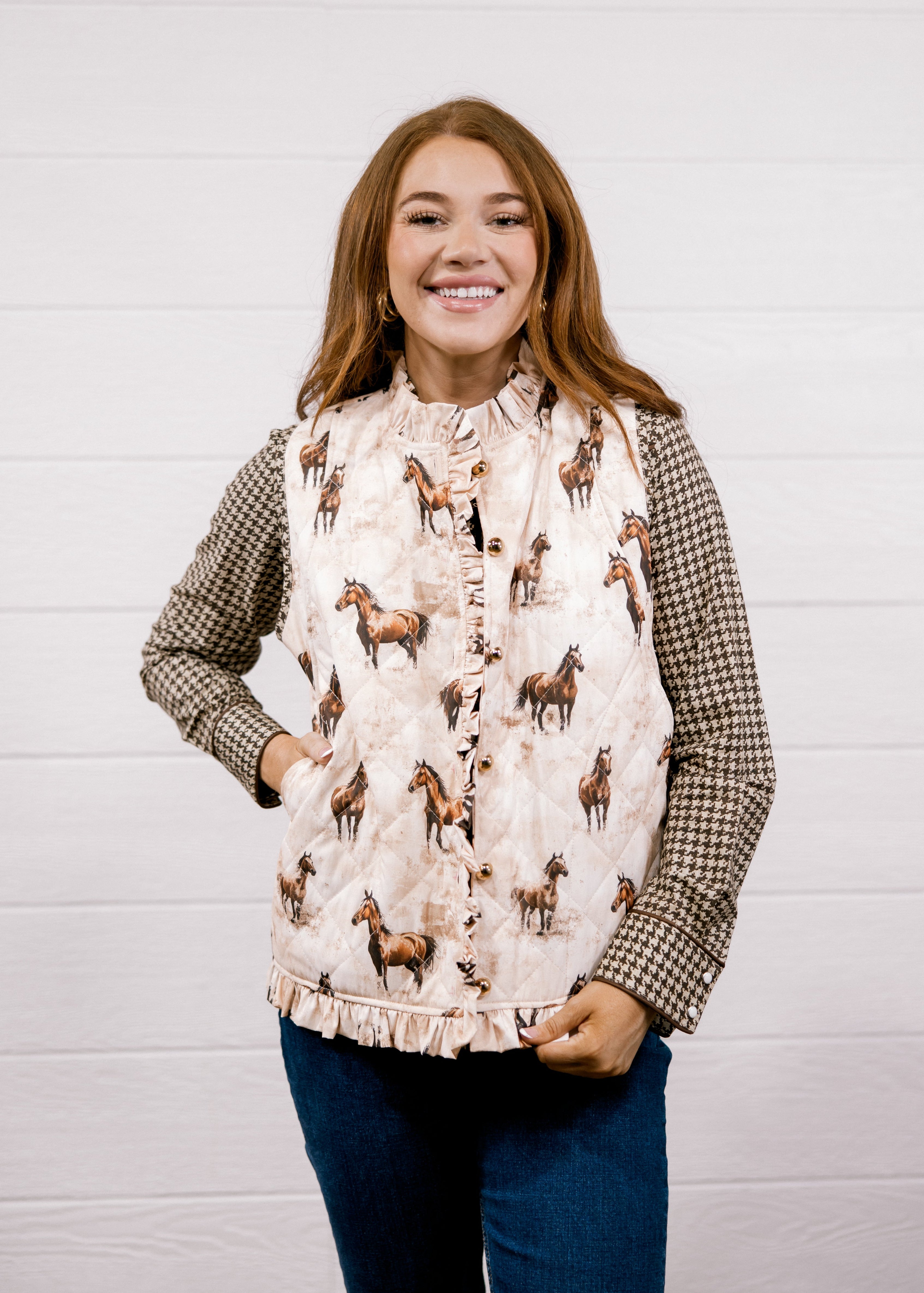 A smiling woman wearing a sleeveless western-style vest with a beige and brown horse print over a brown-and-white houndstooth shirt, paired with dark blue jeans, standing against a white shiplap wall.