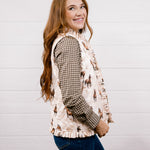 A woman with long light brown hair is shown from the waist up, wearing a cream-colored western-style vest with a horse pattern over a brown and black houndstooth shirt, paired with blue jeans, against a white shiplap wall.
