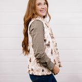 A woman with long light brown hair is shown from the waist up, wearing a cream-colored western-style vest with a horse pattern over a brown and black houndstooth shirt, paired with blue jeans, against a white shiplap wall.