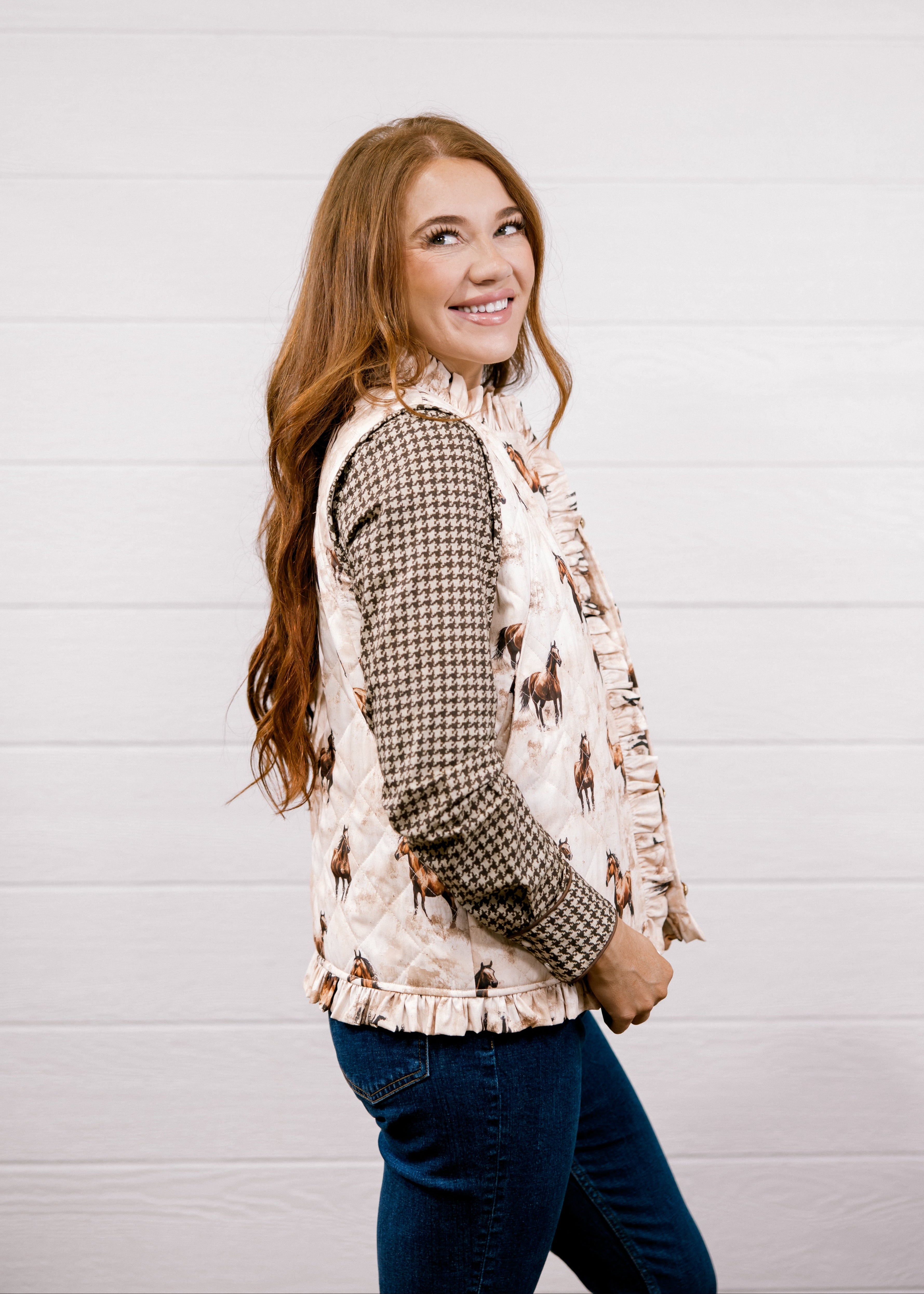 A woman with long light brown hair is shown from the waist up, wearing a cream-colored western-style vest with a horse pattern over a brown and black houndstooth shirt, paired with blue jeans, against a white shiplap wall.