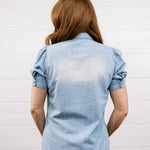 A woman viewed from behind wearing a light blue chambray short-sleeve button-up shirt and dark blue jeans, standing against a white paneled wall.