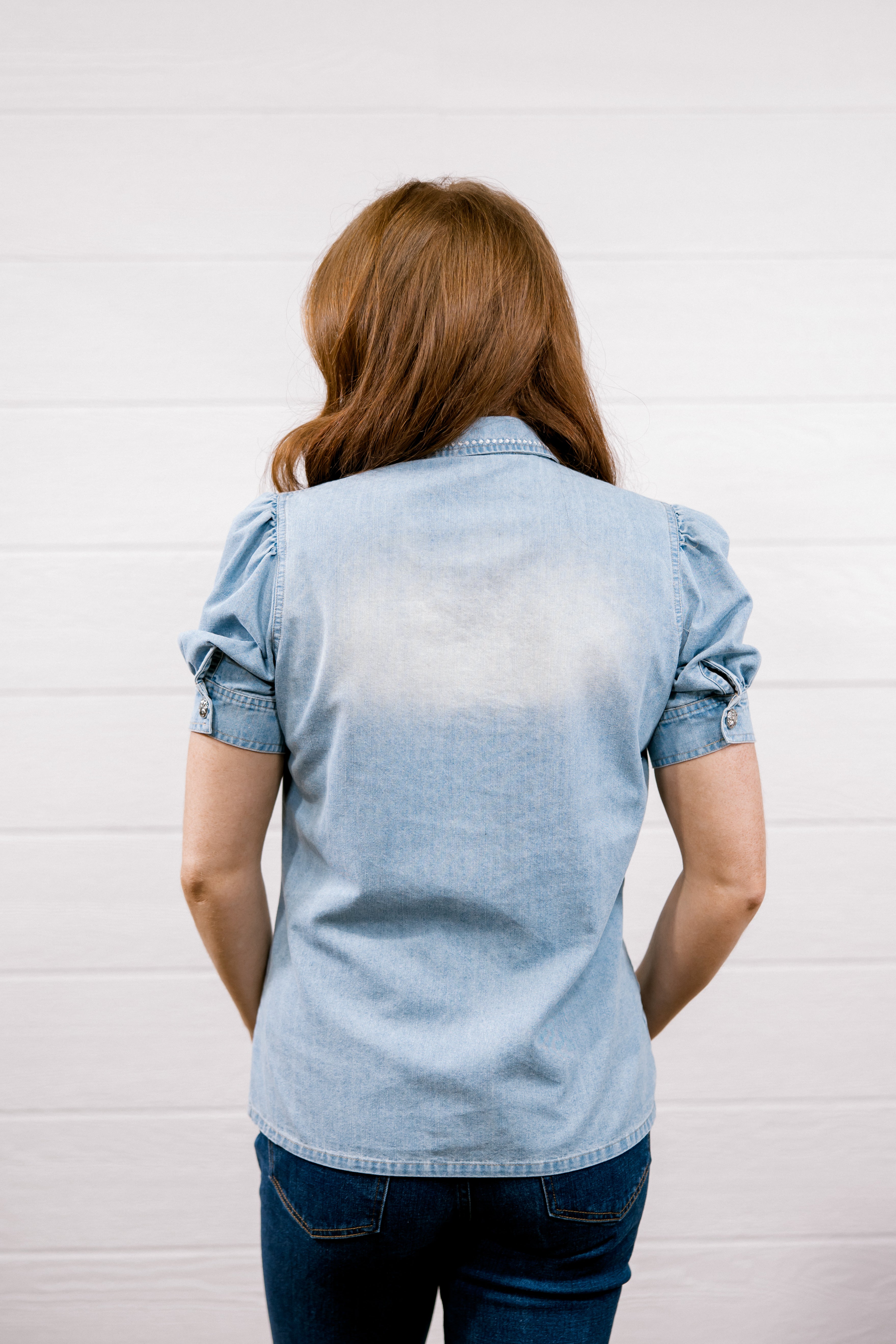 A woman viewed from behind wearing a light blue chambray short-sleeve button-up shirt and dark blue jeans, standing against a white paneled wall.