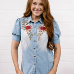 A smiling woman with long red hair wearing a light blue denim shirt with red and gold floral embroidery on the chest, paired with dark blue jeans, standing against a white shiplap wall.