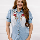 A smiling woman with long red hair wearing a light blue denim shirt with red and gold floral embroidery on the chest, paired with dark blue jeans, standing against a white shiplap wall.