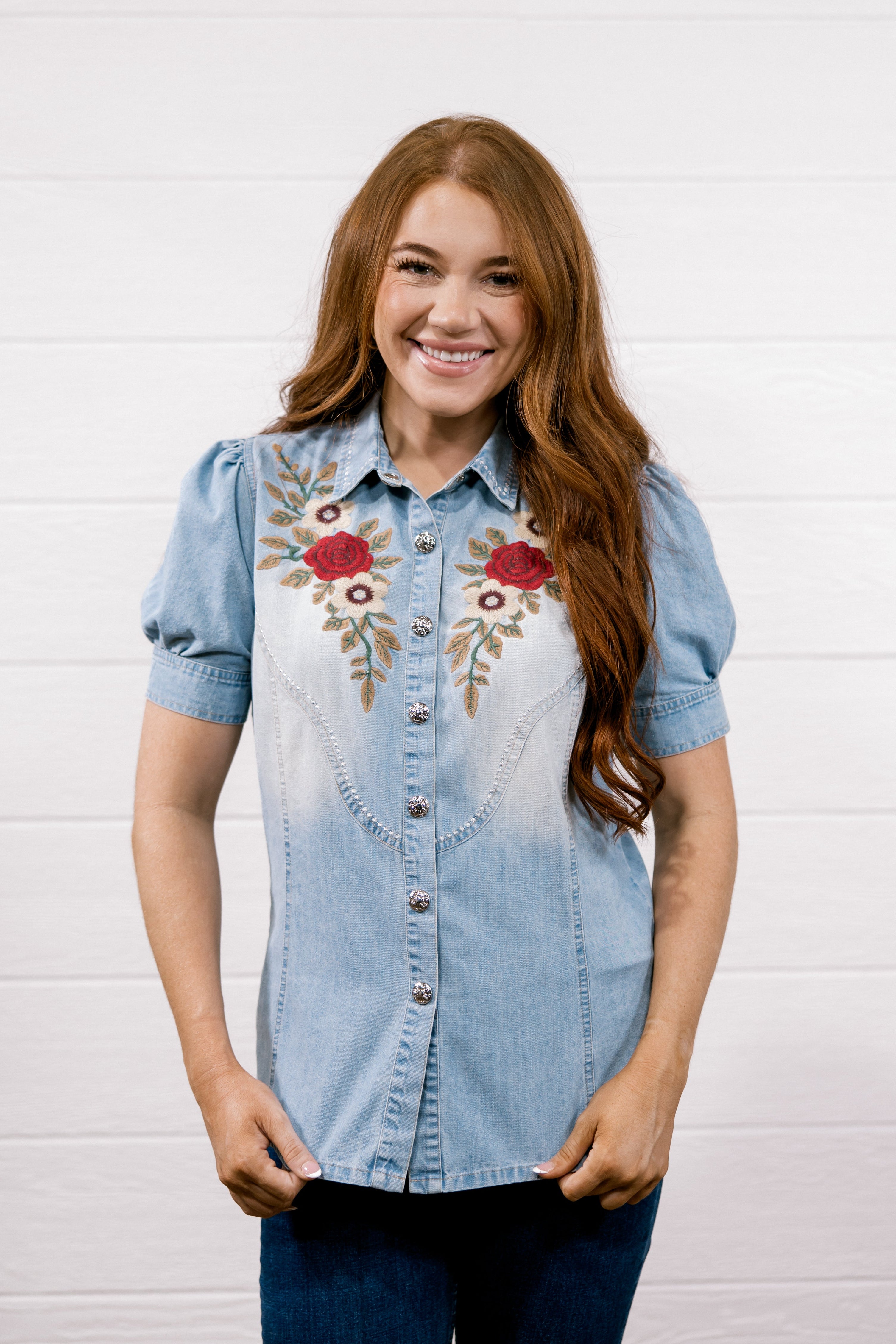 A smiling woman with long red hair wearing a light blue denim shirt with red and gold floral embroidery on the chest, paired with dark blue jeans, standing against a white shiplap wall.