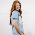 A smiling young woman with long light brown hair is shown in profile wearing a light-wash blue denim shirt and dark blue jeans, standing against a white wood-paneled wall.