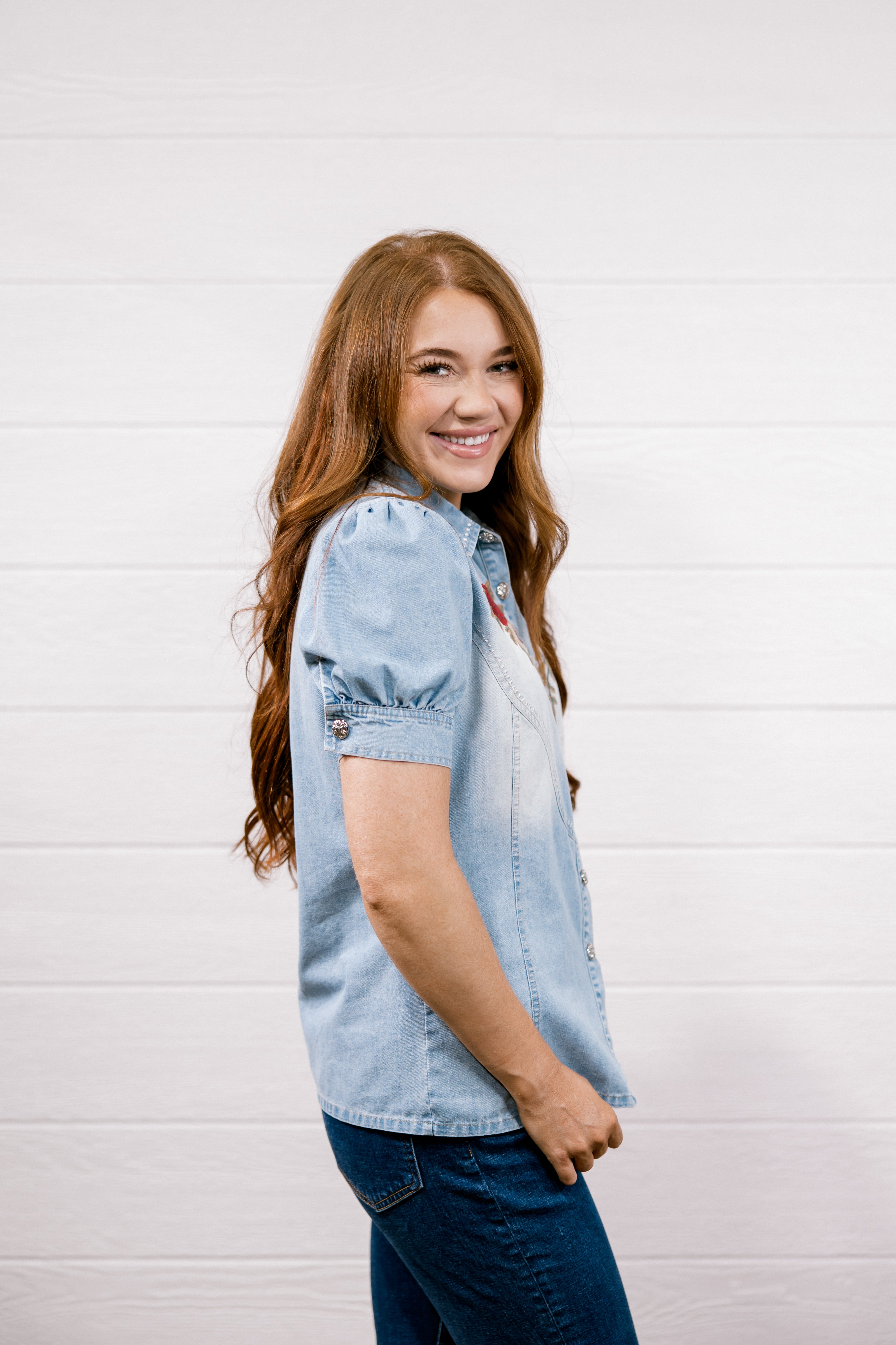A smiling young woman with long light brown hair is shown in profile wearing a light-wash blue denim shirt and dark blue jeans, standing against a white wood-paneled wall.