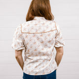 A woman viewed from behind wearing a short-sleeve button-up shirt with a small floral pattern and contrast piping, paired with dark blue jeans, standing against a white paneled wall.