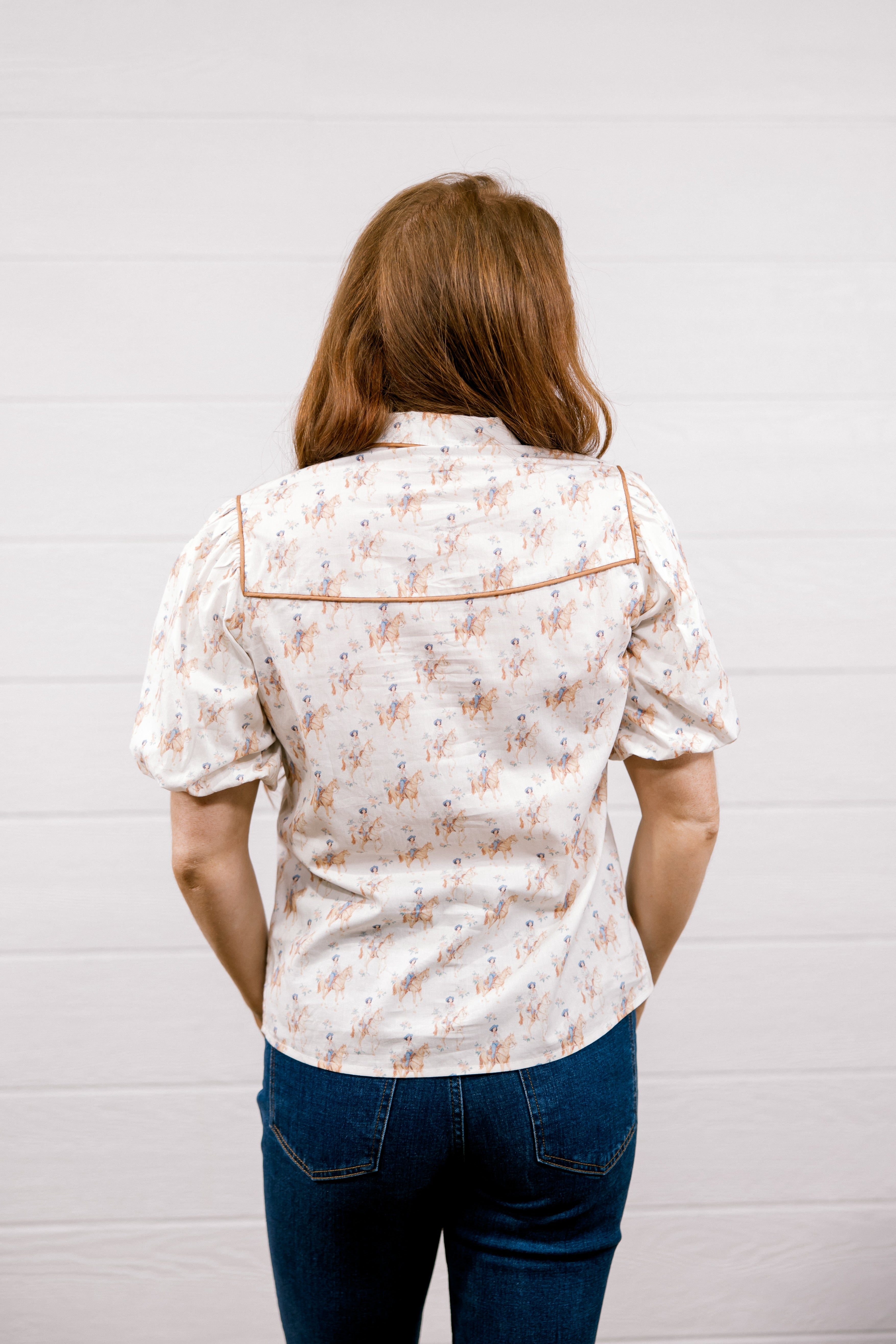 A woman viewed from behind wearing a short-sleeve button-up shirt with a small floral pattern and contrast piping, paired with dark blue jeans, standing against a white paneled wall.