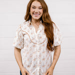 A smiling young woman with long wavy light brown hair stands against a white shiplap wall, wearing a short-sleeve button-up blouse with a beige and orange floral pattern and dark blue jeans.