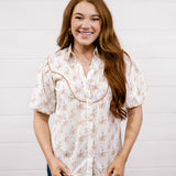 A smiling young woman with long wavy light brown hair stands against a white shiplap wall, wearing a short-sleeve button-up blouse with a beige and orange floral pattern and dark blue jeans.