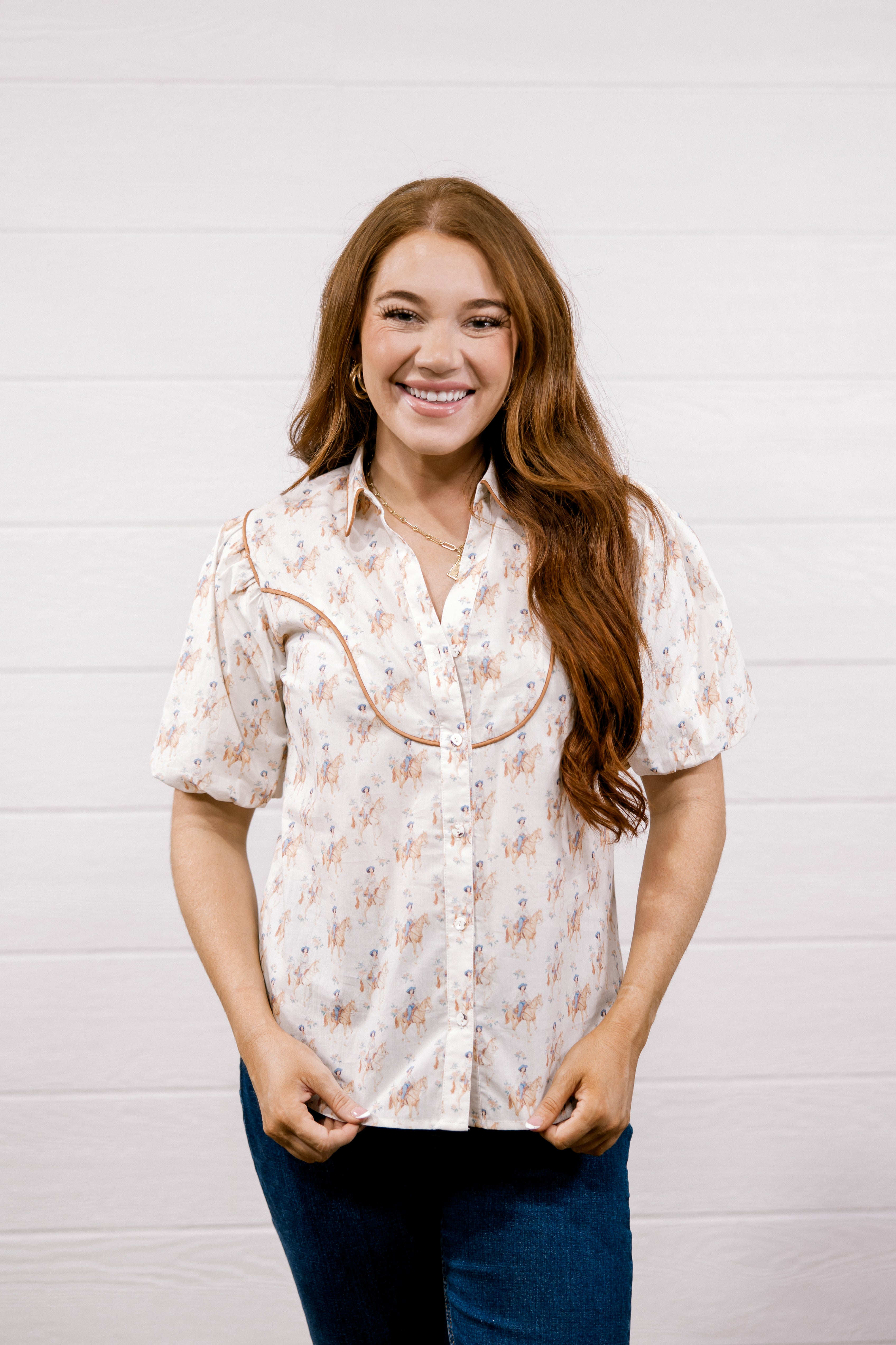 A smiling young woman with long wavy light brown hair stands against a white shiplap wall, wearing a short-sleeve button-up blouse with a beige and orange floral pattern and dark blue jeans.