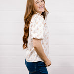 A woman with long wavy light brown hair is shown from the side, wearing a short-sleeve button-up shirt with a delicate cowgirl pattern, paired with dark blue jeans, against a white shiplap wall background.