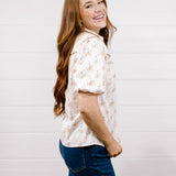 A woman with long wavy light brown hair is shown from the side, wearing a short-sleeve button-up shirt with a delicate cowgirl pattern, paired with dark blue jeans, against a white shiplap wall background.