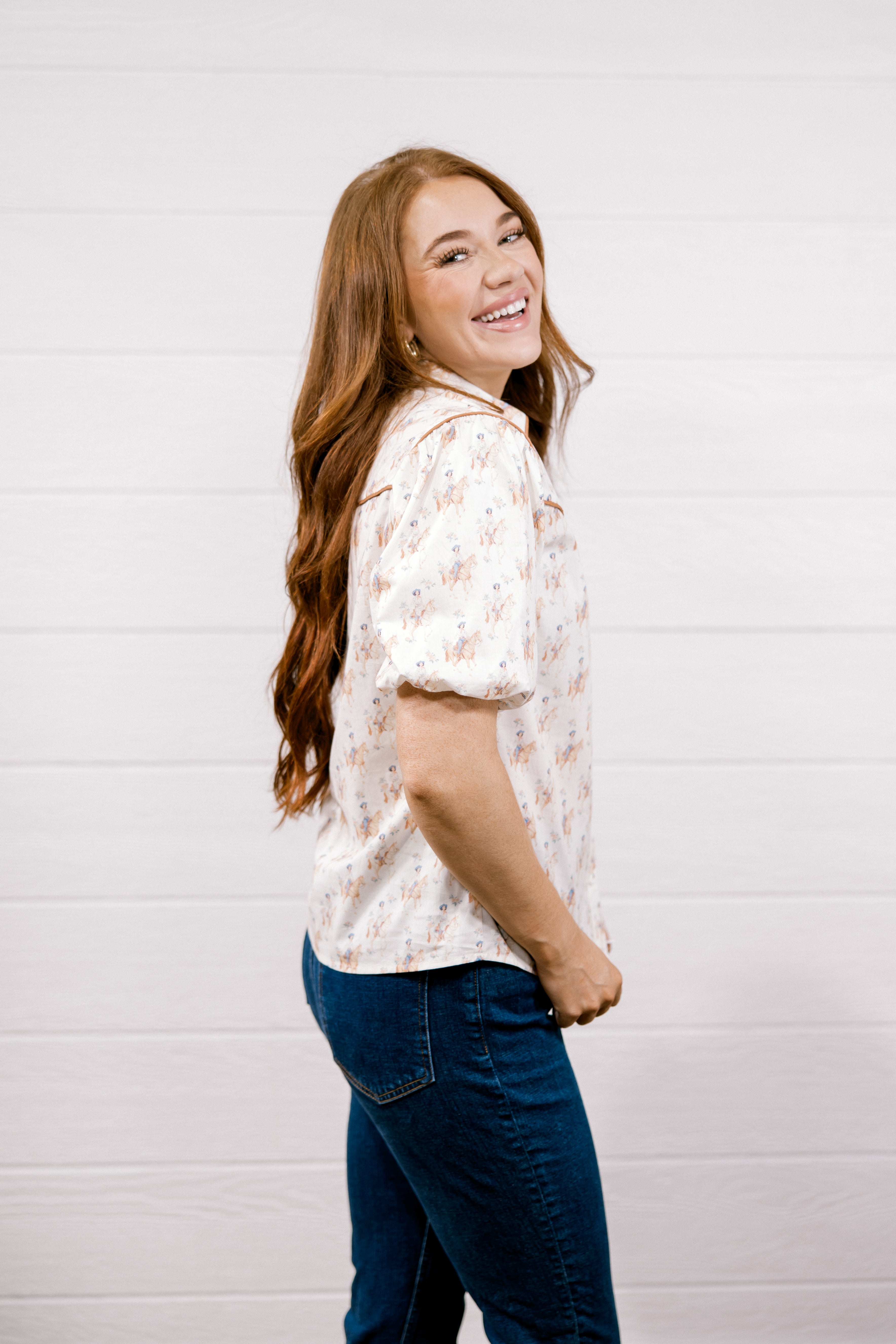 A woman with long wavy light brown hair is shown from the side, wearing a short-sleeve button-up shirt with a delicate cowgirl pattern, paired with dark blue jeans, against a white shiplap wall background.