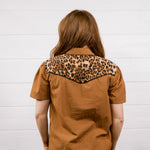 A woman viewed from behind wearing a short-sleeve brown shirt with a leopard-print yoke across the shoulders, paired with blue jeans, standing against a white paneled wall.
