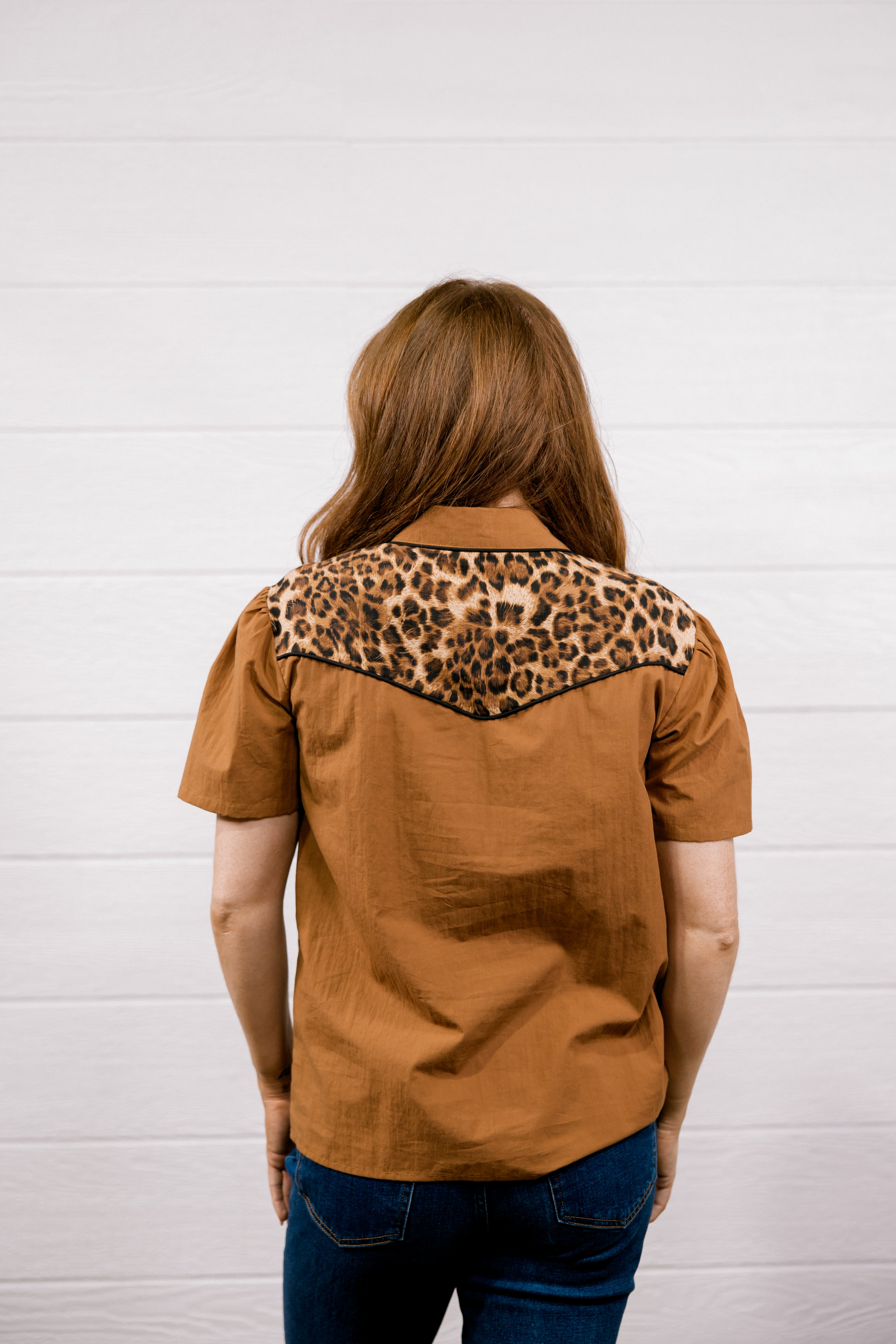 A woman viewed from behind wearing a short-sleeve brown shirt with a leopard-print yoke across the shoulders, paired with blue jeans, standing against a white paneled wall.