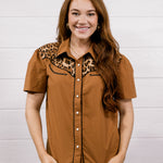 A woman modeling a short-sleeve button-up shirt in a warm brown color with a leopard print collar, paired with blue jeans, standing against a white wood-paneled wall.