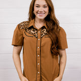 A woman modeling a short-sleeve button-up shirt in a warm brown color with a leopard print collar, paired with blue jeans, standing against a white wood-paneled wall.