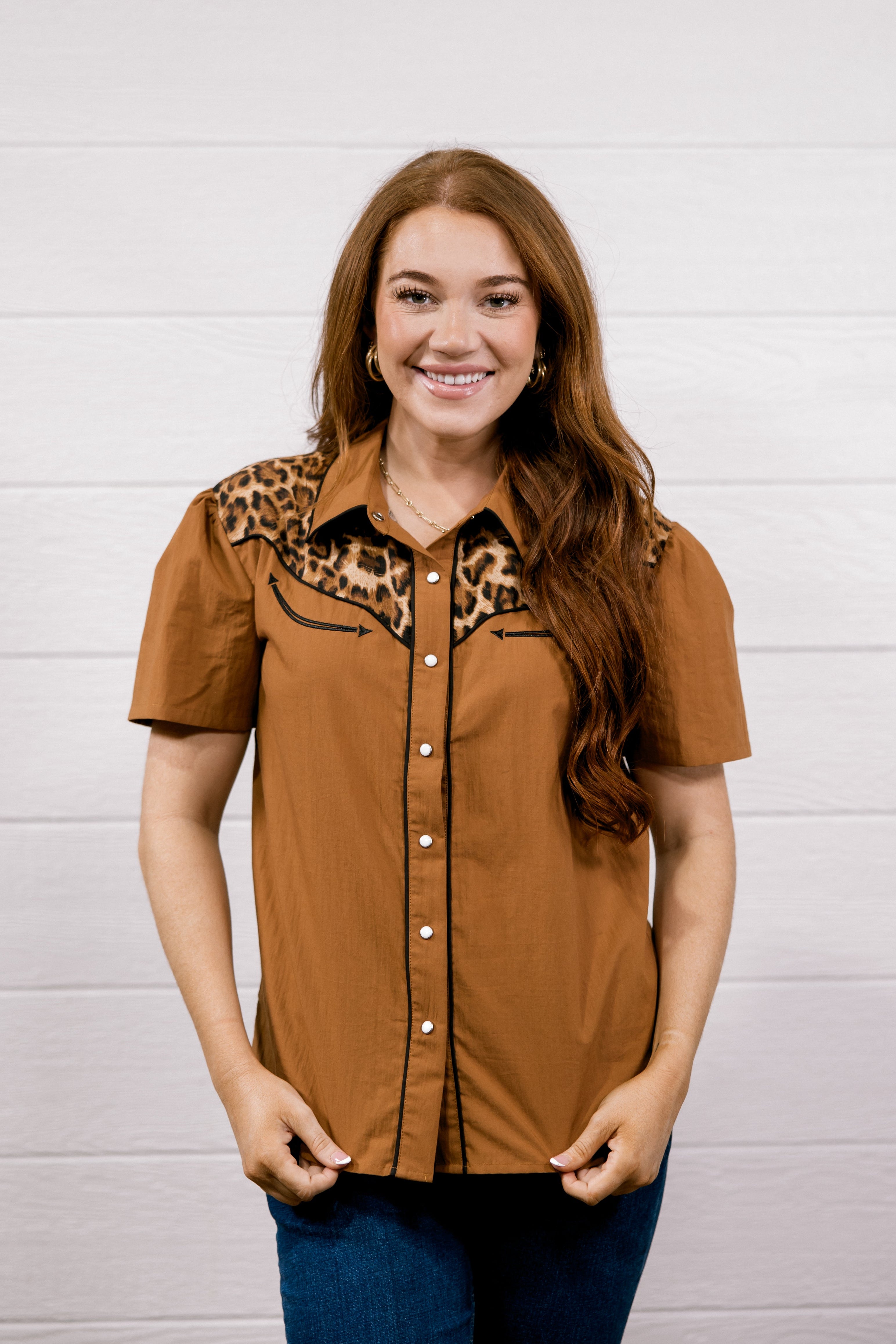 A woman modeling a short-sleeve button-up shirt in a warm brown color with a leopard print collar, paired with blue jeans, standing against a white wood-paneled wall.