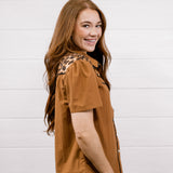A woman with long light brown hair is shown from the waist up, wearing a short-sleeve button-up shirt in a warm camel brown color with a leopard print pattern on the shoulders, paired with dark blue jeans. She is smiling and looking over her shoulder against a white wood-paneled wall.