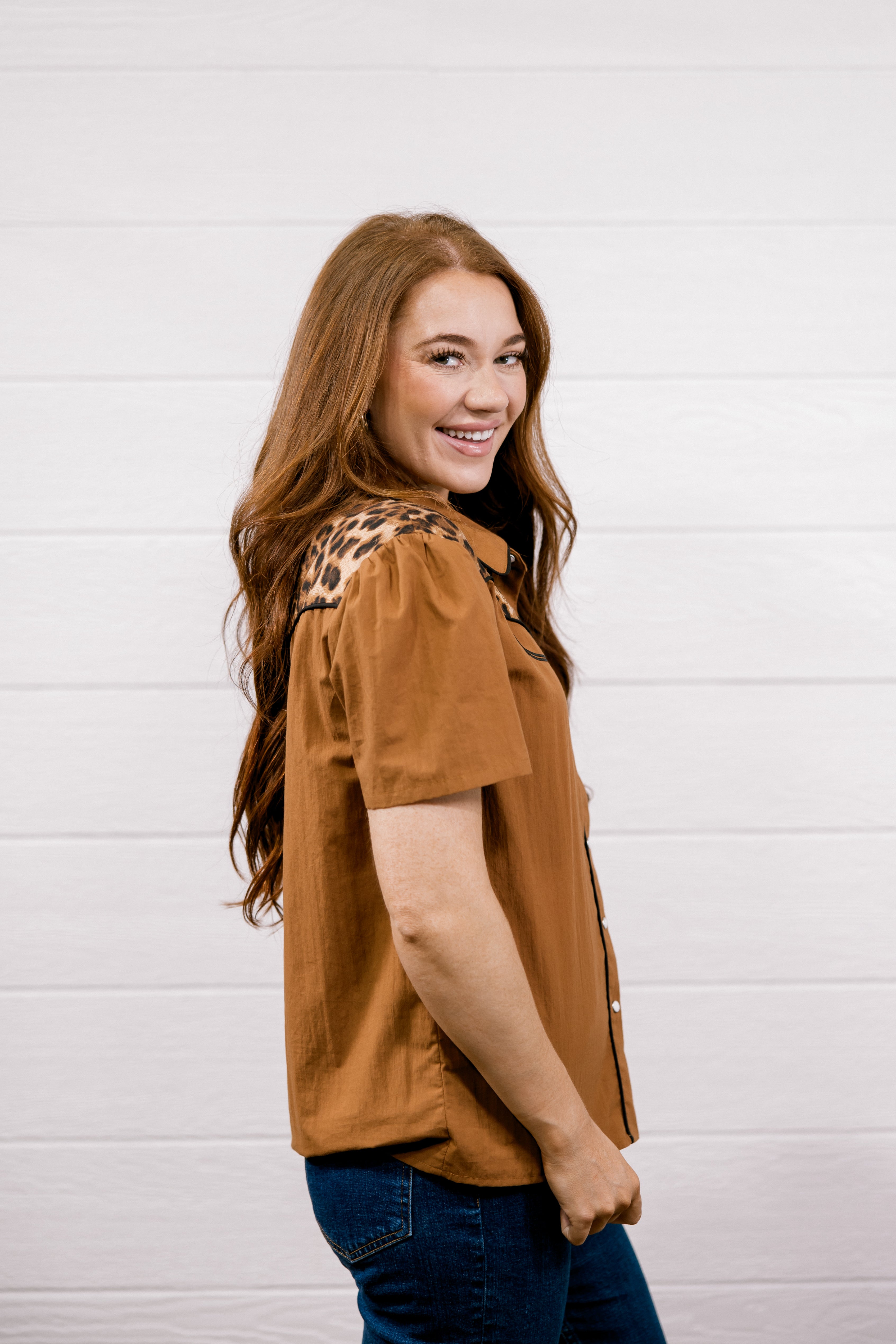 A woman with long light brown hair is shown from the waist up, wearing a short-sleeve button-up shirt in a warm camel brown color with a leopard print pattern on the shoulders, paired with dark blue jeans. She is smiling and looking over her shoulder against a white wood-paneled wall.
